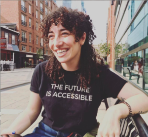 Rachele Manett smiling while sitting on a bench. Rachelle is wearing a black t-shirt that says, the future is accessible.