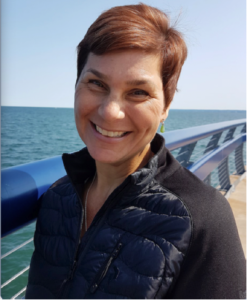 a woman with dark brown hair, on a boat, with blue skies in behind, she is smiling at the camera