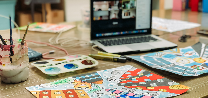 A laptop is open and shows a zoom screen at the top right of this image. In the foreground, various art supplies and a project are placed on a table.