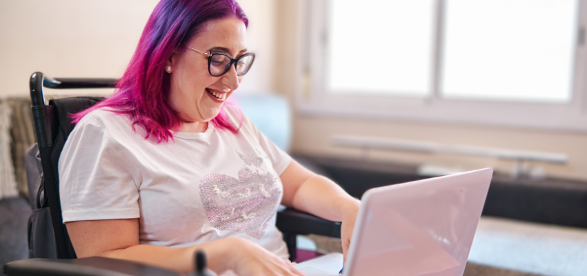 A woman with pink and purple hair wears glasses and sits in a wheelchair. She smiles as she looks at her laptop.